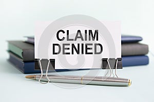 a white card with claim denied text stands on paper clips on a desk against the light blue background, selective focus