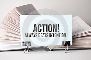 A white card with the text Action Always beats intention stands on a clip for papers on the table against the background of books