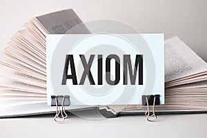 A white card with the text axiom stands on a clip for papers on the table against the background of books. Defocus