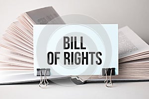 A white card with the text Bill of Rights stands on a clip for papers on the table against the background of books. Defocus