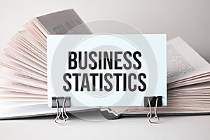 A white card with the text business statistics stands on a clip for papers on the table against the background of books. Defocus