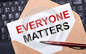 White card with the text EVERYONE MATTERS in a craft envelope on a work desk with a modern laptop keyboard and burgundy pen. Flat