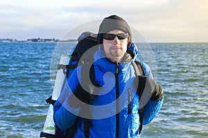 White Caucasian male traveler in a sports blue jacket, gloves, hat, glasses and a backpack stands against the background