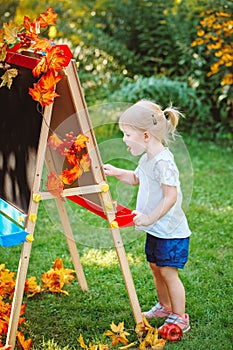 White Caucasian toddler child kid girl standing outside in summer autumn park drawing on easel with markers