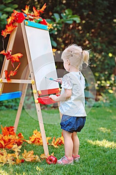 White Caucasian toddler child kid girl standing outside in summer autumn park drawing on easel with markers