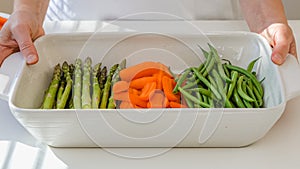 White ceramic baking dish, and fresh vegetables, carrot, green beans, asparagus. close up on white kitchen table. Cooking process
