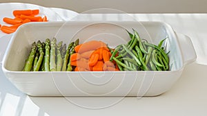 White ceramic baking dish, and fresh vegetables, carrot, green beans, asparagus. close up on white kitchen table. Cooking process