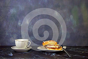 A white ceramic cup with black coffee and a plate with two cinnamon rolls in icing and a fork on the marble table
