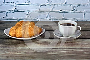 White ceramic cup with black coffee, a spoon and a plate with two fresh croissants