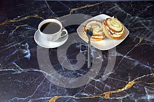 A white ceramic cup of coffee on a saucer and a plate with two cinnamon rolls in a glaze and a fork on a marble table