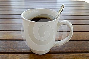 A white ceramic cup with a morning refreshing hot coffee with tea drink and tea shiny teaspoon is on a wooden table in a cafe