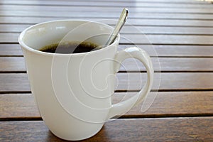 A white ceramic cup with a morning refreshing hot coffee with tea drink and tea shiny teaspoon is on a wooden table in a cafe