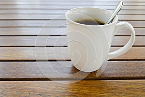 A white ceramic cup with a morning refreshing hot coffee with tea drink and tea shiny teaspoon is on a wooden table in a cafe