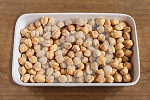 A white ceramic dish full of raw white chickpeas with a yellowish tone after being soaked on a wooden table.