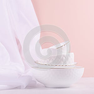White ceramic dishes pile with thin gold border with airy silk curtain on white wood table in delicate pastel pink interior.