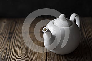 White ceramic mockup teapot for hot drinks on empty old wooden table near black wall in evening light with shadows