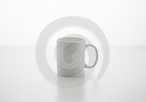 White ceramic mug with a glossy finish on a light-colored, smooth surface. The mug is