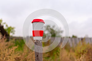 A white ceramic mug with a red lid stands with a drink on a wooden pole in the village in autumn.