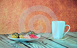 A white ceramic mug with tea and a plate with three eclairs and a red flower stand on a wooden table