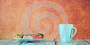 A white ceramic mug with tea and a plate with three eclairs and a red flower stand on a wooden table