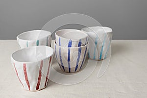 White ceramic mugs with colorful stripes on table with linen tablecloth and gray wall background. Handmade