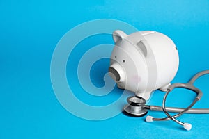 White ceramic piggy bank and stethoscope on light blue background, space for text. Medical insurance