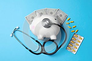 White ceramic piggy bank, stethoscope, money and pills on light blue background, flat lay. Medical insurance