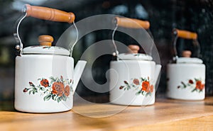 White ceramic tea pots with wooden handle, painted with red roses and displayed on a wooden shelf