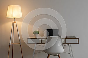 White chair, laptop with blank screen, potted plant on table and glowing lamp in evening on gray wall background
