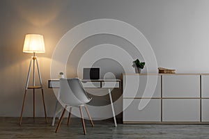 White chair, laptop with blank screen on table, glowing lamp and plant in pot in evening on gray wall background