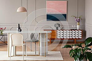 White chair and marble table under pink lamp in eclectic living room interior with painting above cabinet. Real photo