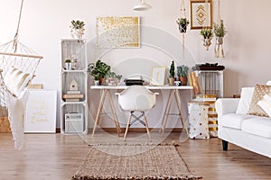 White chair placed by the desk with plants and typewriter in real photo of bright living room interior with posters and rug