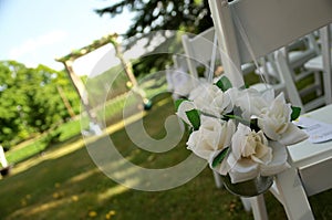 White Chairs Adorned with White Fabric Rose Bouquets Await Guests at a Garden Wedding