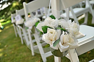 White Chairs Adorned with White Fabric Rose Bouquets Await Guests at a Garden Wedding