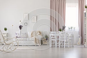 White chairs at dining table in apartment interior with blanket on sofa and pink drapes. Real photo