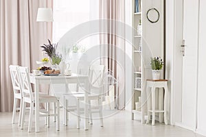 White chairs at dining table with plants in modern flat interior with pink drapes and window. Real photo