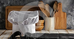 White chef`s hat and wooden table background with dark wall and kitchen utensils.