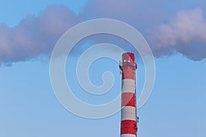 White chimney of a factory, from which gray smoke comes out against a blue sky on a sunny frosty winter day