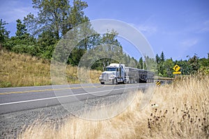 White classic big rig semi truck transporting cargo in covered bulk semi trailer running on the road with hills on the sides in