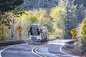 White classic car hauler big rig semi truck transporting cars on hydraulic semi trailer driving on the winding road going through