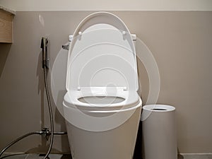 White clean ceramic toilet bowl, front view with silver bidet shower or health faucet beside the bin in modern bathroom.