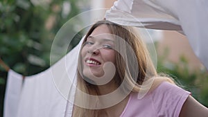 White clean laundry hanging on rope outdoors with cheerful young Caucasian woman peeking smiling looking at camera