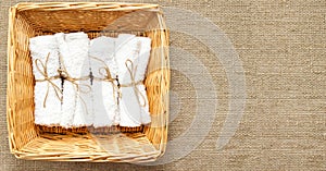 White clean rolled terry towel stack in wicker basket on natural linen sack background. Flat Lay