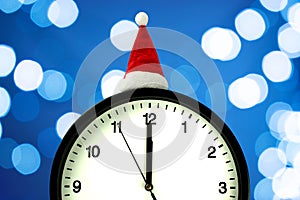 The white clock in red sunta hat shows five to seconds midnight, on the blurry background. Countdown to the new year