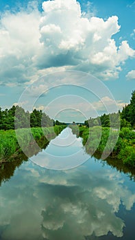 White clouds and blue sky are reflected in the calm water of the river. Trees and shrubs grow along the irrigation canal. Autumn