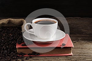 A white coffee cup with black coffee is placed on a red book and a large number of roasted coffee beans are placed around on a