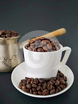 White coffee cup and saucer; coffee beans scattered on the table. In the background is a cezve. Black background.