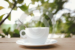 A white coffee cup with spoon in a cup and saucer are placed on a wooden plate and on the landscape nature background