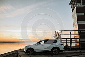 White compact SUV car with sport and modern design parked on concrete road by the sea near the beach at sunset