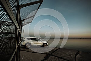 White compact SUV car with sport and modern design parked on concrete road by the sea near the beach at sunset